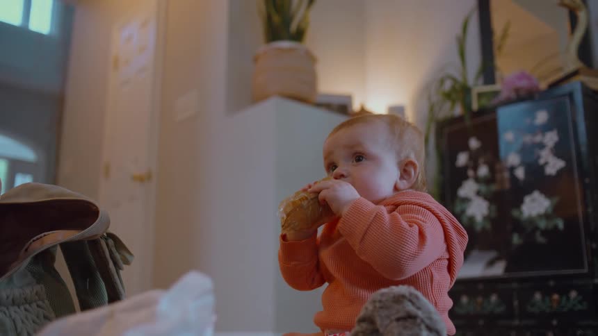 A cute baby girl in an orange hoodie sits on the floor, receives a pastry, and attempts to eat it