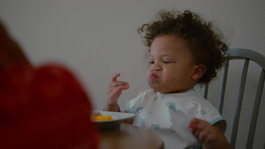 An adorable toddler with curly hair smiles and makes funny faces while eating food from a plate in a high chair