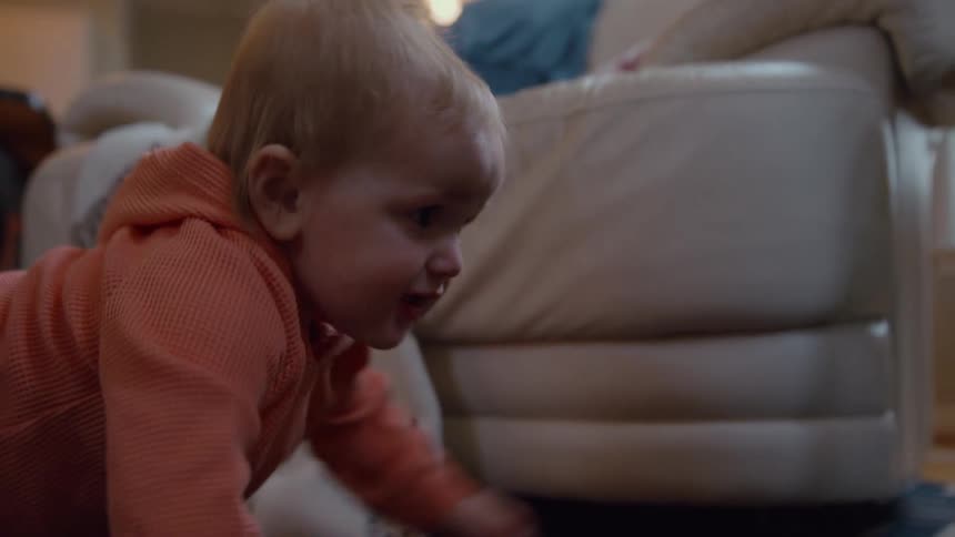 A joyful red-haired baby in an orange outfit laughs and crawls across a patterned rug in a cozy living room in slow motion