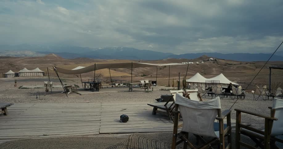 A large white sunshade canopy billows in the wind at a luxury desert campsite with mountains in the background under a cloudy sky