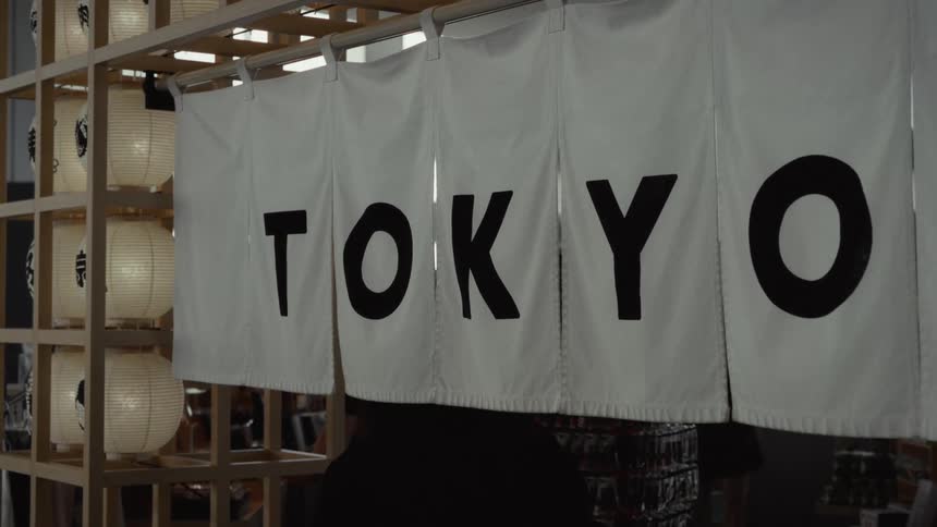 A white noren curtain with 'Tokyo' in black letters hangs in a modern Japanese-style setting with paper lanterns