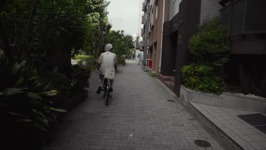 A slow-motion shot follows an elderly woman as she calmly rides her bicycle down a narrow brick alley in a quiet Tokyo neighborhood