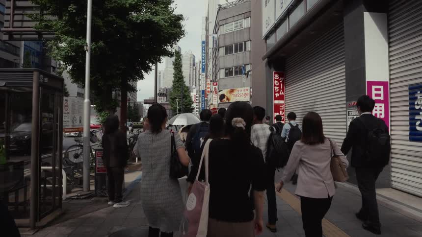 A slow-motion point-of-view shot follows a crowd of pedestrians walking down a busy city street in Shinjuku, Tokyo