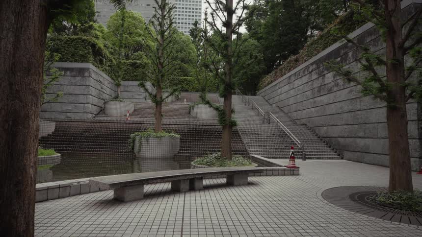 A slow pull-back shot reveals a serene, modern urban park with geometric concrete architecture, trees, and a tranquil water feature