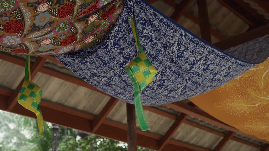 Traditional green and yellow ketupat decorations sway gently from a ceiling adorned with colorful patterned fabrics