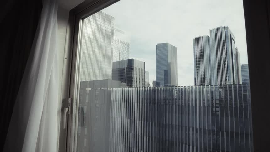 A slow push-in shot reveals a modern cityscape of glass skyscrapers through a room window on an overcast day