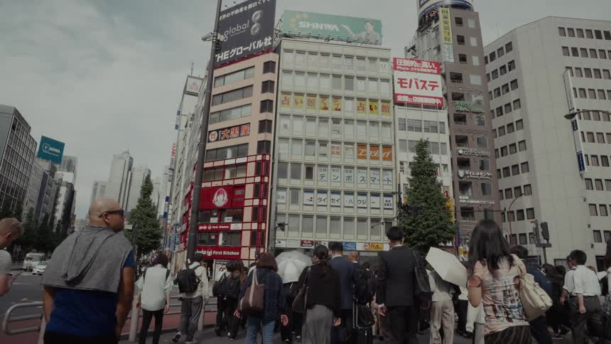 A diverse crowd of people walks in slow motion across a busy street in a dense urban city in Tokyo, Japan