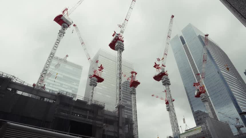 A low-angle view of several large construction cranes towering over a building site in a modern city on a cloudy day