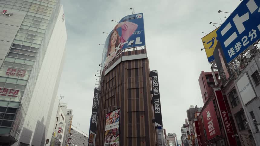 A low-angle shot captures the modern architecture and vibrant billboards of Paselabo Tower in a Tokyo city street