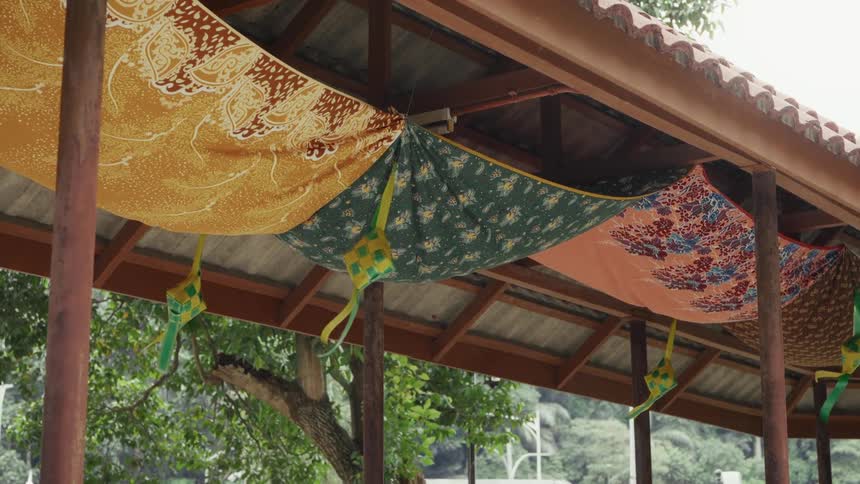 Traditional Malay Hari Raya decorations, featuring colorful fabrics and woven ketupat, adorn the ceiling of an outdoor pavilion