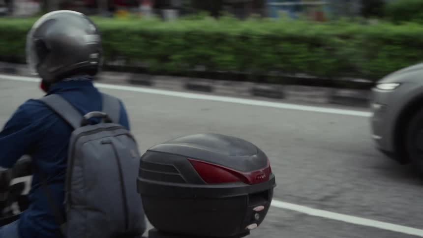 A man on a scooter with a backpack drives through city traffic on a paved road during the day