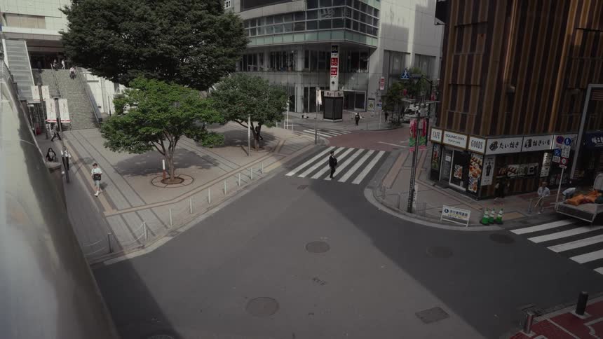 Pedestrians and a small truck cross a sunlit street corner in a modern Japanese city in slow motion