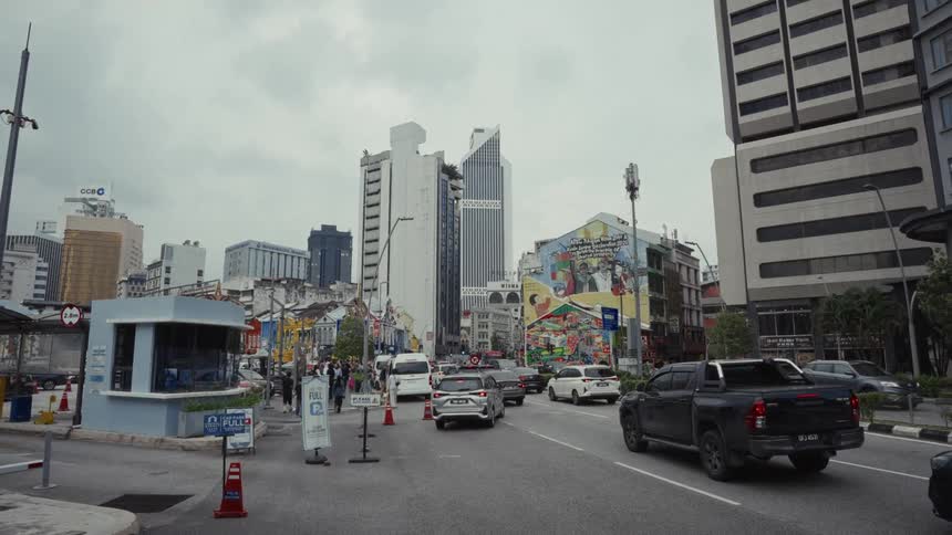 An establishing shot of a city street in Kuala Lumpur shows traffic and pedestrians with a large, colorful mural in the background on a cloudy day