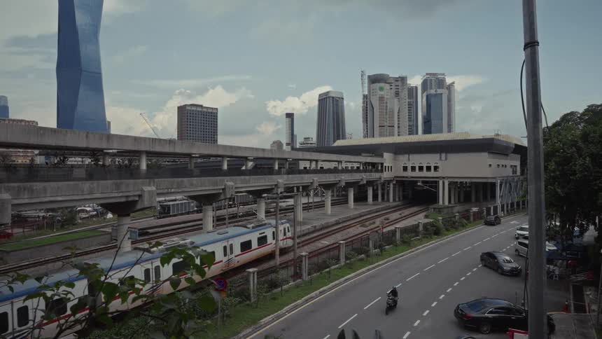 A commuter train departs from a busy urban station, with the modern city skyline of Kuala Lumpur visible in the background under a cloudy sky