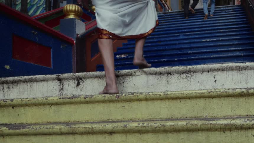 A person in traditional clothing walks barefoot up the colorful weathered steps of a temple in slow motion