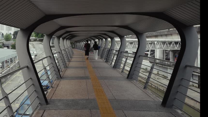 A wide shot shows two people walking away from the camera down a long, modern covered pedestrian skybridge