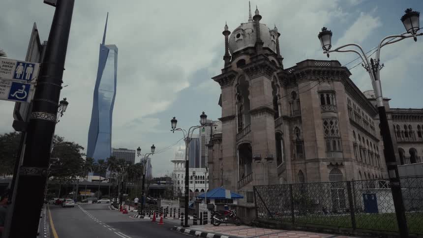 A historic government building stands next to an almost empty road with the modern Merdeka 118 tower in the background on a cloudy day in Kuala Lumpur