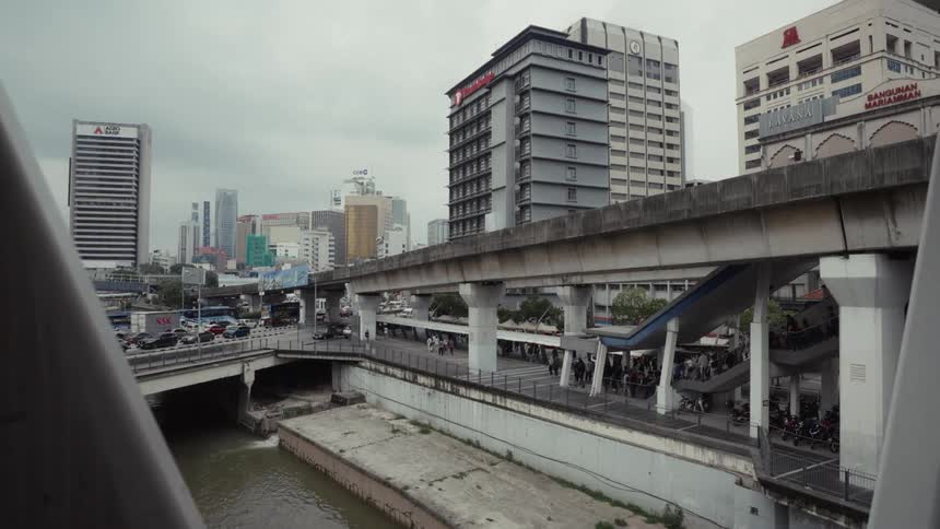 A slow zoom out reveals the urban landscape of Kuala Lumpur with traffic, a river, and an elevated train station under an overcast sky