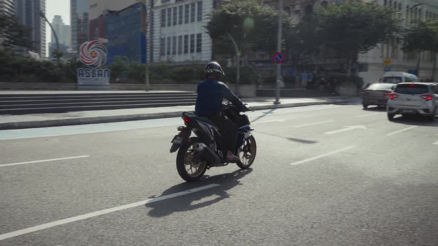 A man wearing a helmet rides a motorcycle down a sunny city street in slow motion
