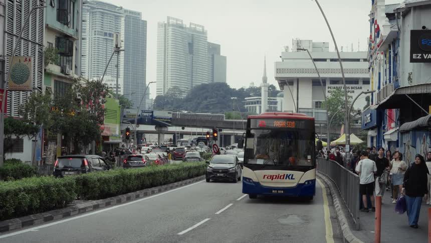 A public transit bus slowly drives through a bustling street in downtown Kuala Lumpur as pedestrians walk along the sidewalk