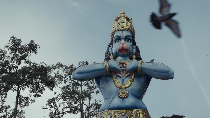 A majestic blue statue of the Hindu God Hanuman stands tall as a bird flies by in slow motion