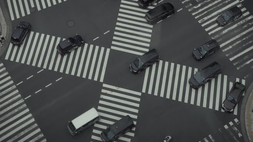 Birds-eye view of cars, trucks, and motorcycles methodically crossing a busy urban intersection with striking white crosswalks