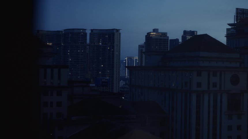 A still cityscape of tall buildings with lights on stands against the deep blue sky at dusk