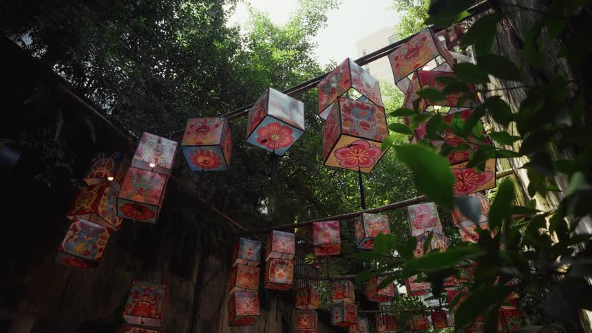 Vibrant cube-shaped lanterns with traditional Asian art hang in a lush, green alleyway during a cultural festival