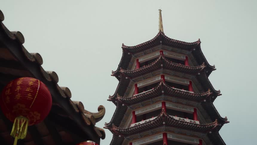 A towering traditional Chinese pagoda stands tall next to a temple roof decorated with red lanterns against a cloudy sky