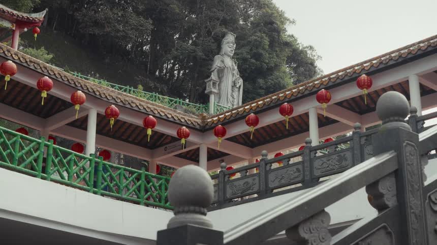 A majestic statue overlooks a covered walkway decorated with red lanterns at a hillside Chinese temple