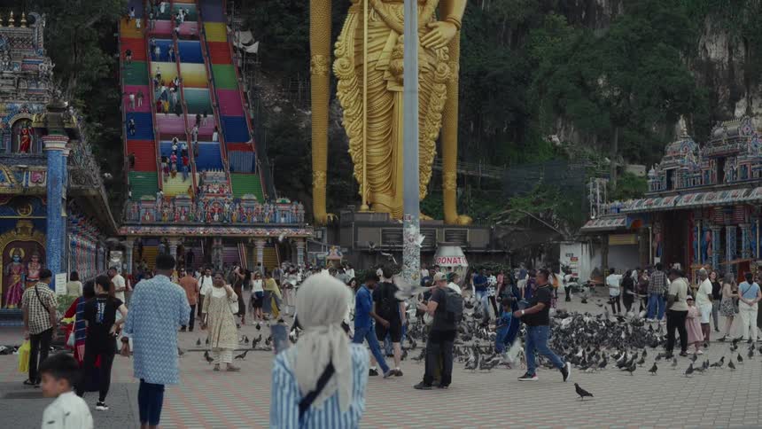A diverse crowd of tourists mills about at the foot of Malaysia's Batu Caves, with pigeons flocking around the giant golden Murugan statue and colorful stairs