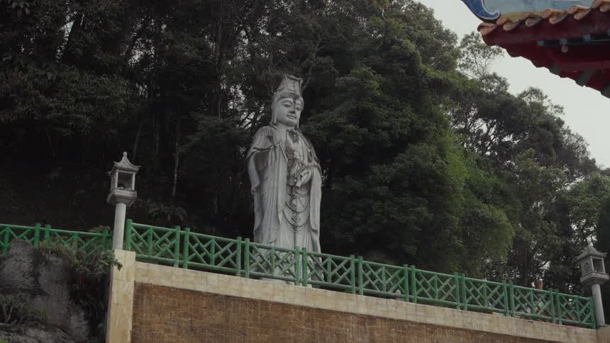 A large stone statue of the Goddess of Mercy, Kuan Yin, stands on a terrace at a mountainside temple as rain falls