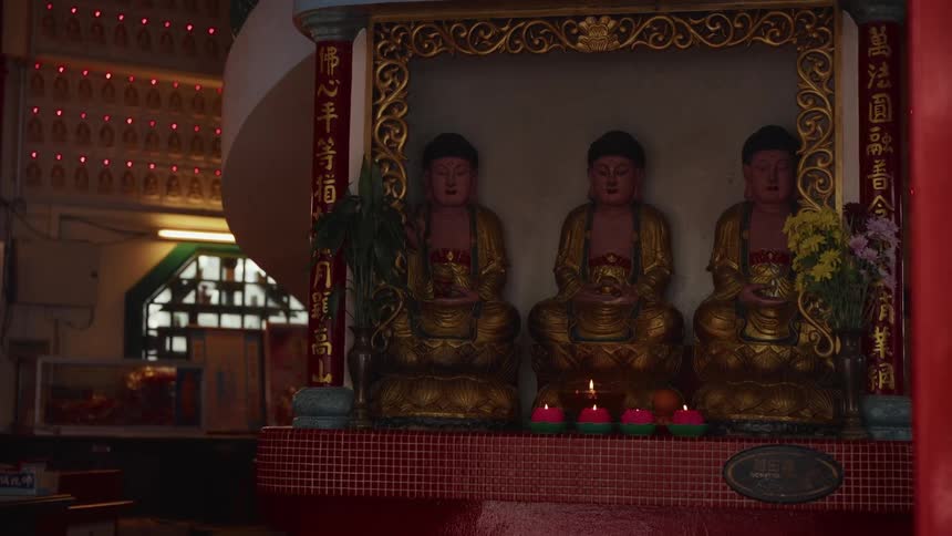 A serene, dimly lit Buddhist shrine features three golden statues with offerings of burning candles and fruit