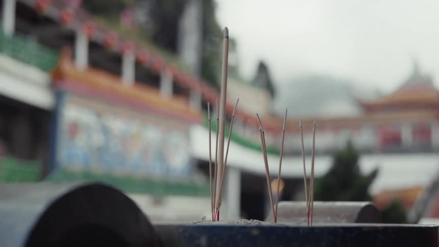 Close up of incense sticks burning in a censer at a Buddhist temple with colorful architecture in the background