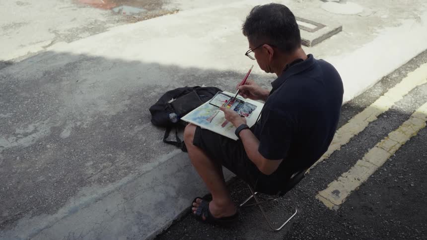 An older Asian artist sits on a folding stool in a shaded urban alley, focused on creating a watercolor painting in his sketchbook