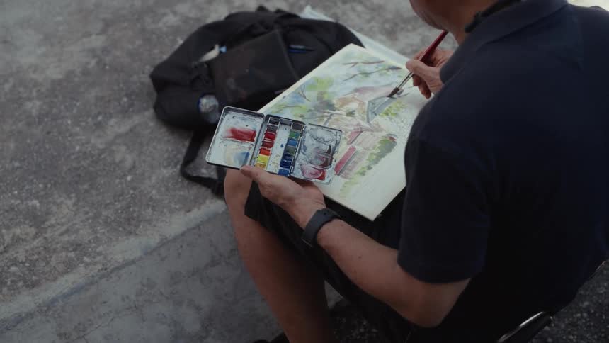 From an over-the-shoulder perspective, an artist carefully applies watercolor paint to a landscape drawing in their sketchbook
