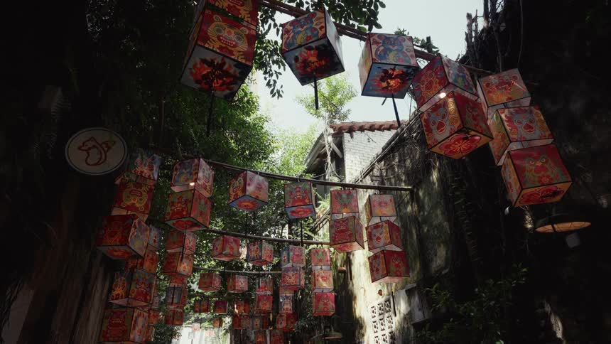 Colorful cube-shaped Chinese lanterns with traditional artwork hang over a narrow, tree-lined alleyway