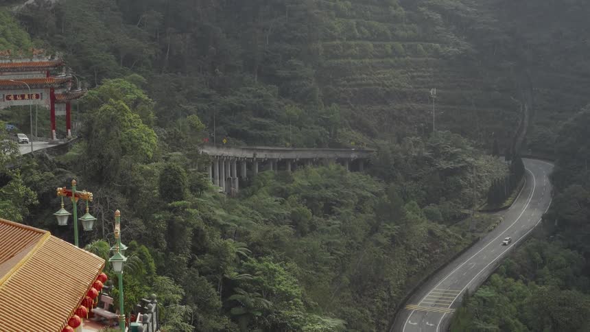 High-angle view of a winding road and bridge through a lush, green mountain valley, with a traditional Asian temple in the foreground