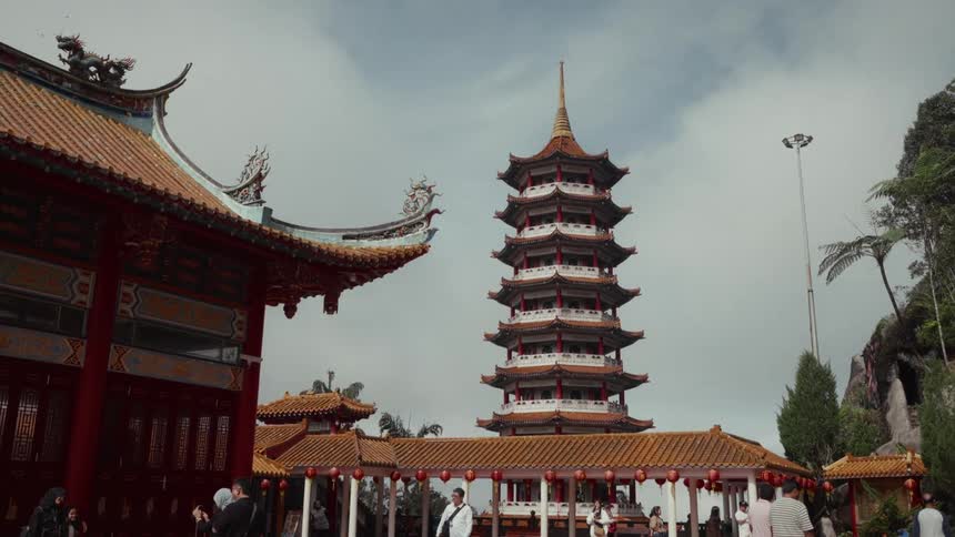 Tourists walk around the Chin Swee Caves Temple grounds with its iconic pagoda towering in the background under a cloudy sky