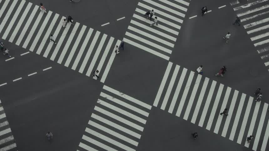 A high-angle, top-down view shows pedestrians slowly crossing a large, striped city intersection