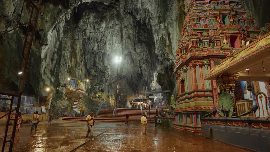 A wide, static shot captures the immense scale of a Hindu temple built inside the vast, illuminated Batu Caves in Malaysia as people walk across the wet floor