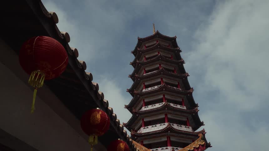 A low-angle view of a majestic Chinese pagoda reaching towards a cloudy sky, with traditional red lanterns in the foreground