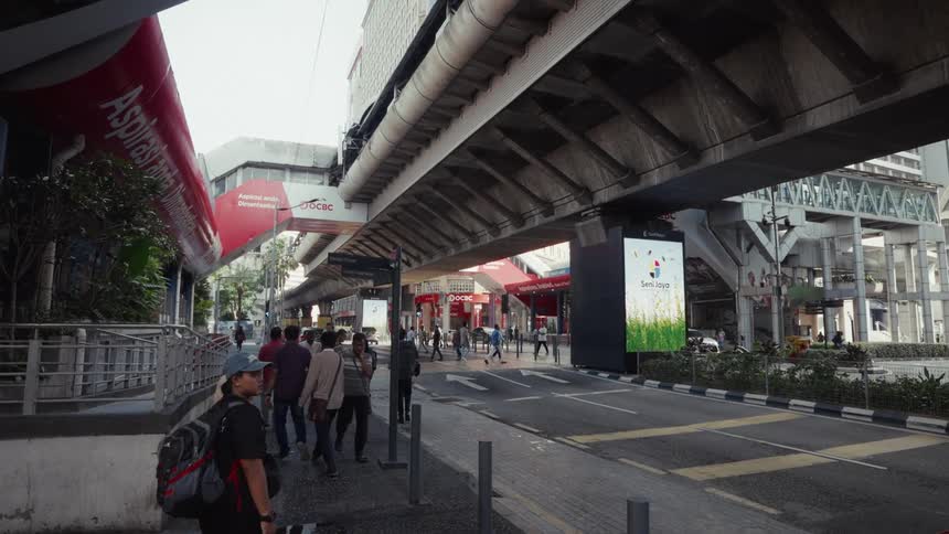 A diverse group of pedestrians walks in slow motion along a sidewalk beneath a large concrete overpass in a bustling modern city