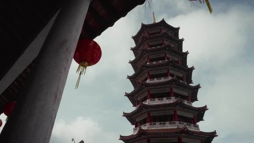 A towering traditional Chinese pagoda stands tall against a cloudy sky, framed by hanging red lanterns