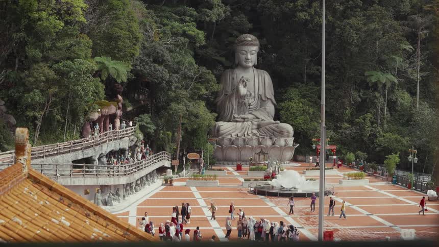 A high-angle view captures tourists wandering through the plaza of a majestic temple with a large seated Buddha statue in a lush forest