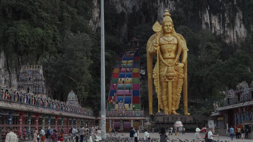 Tourists and worshippers gather at the base of the iconic Batu Caves, featuring the giant golden Lord Murugan statue and the vibrant, colorful staircase in Kuala Lumpur, Malaysia