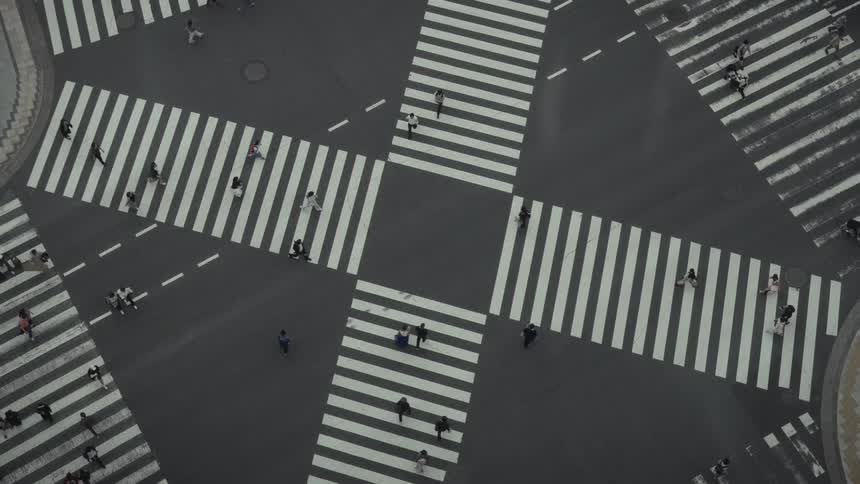 An aerial top-down view captures pedestrians crossing a large, empty city intersection from all directions