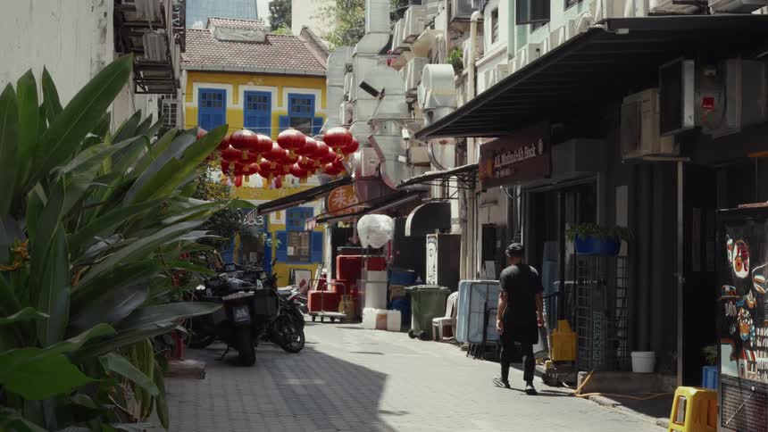 A man walks in slow motion down a vibrant Singapore alley decorated with red Chinese lanterns and lined with colorful buildings