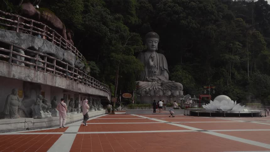 Visitors walk through the plaza of a tranquil mountain temple featuring a giant seated Buddha statue and a lotus fountain