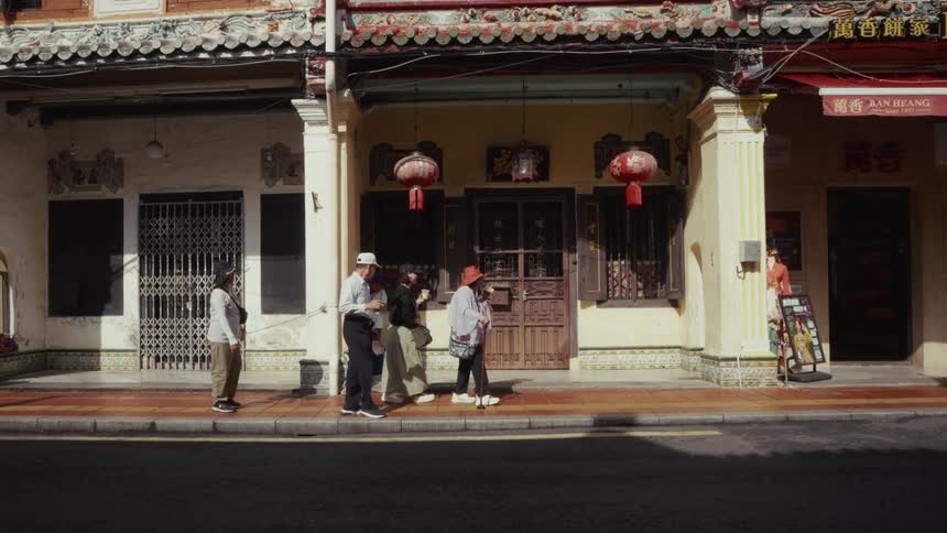 People in slow motion walk along a sunlit sidewalk past ornate and traditional shophouses in an Asian city